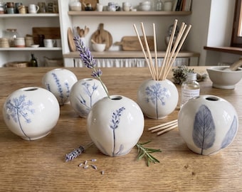 Könnte beinhalten: Ein Set aus fünf kugelförmigen weißen Keramikvasen mit handgemalten blauen botanischen Illustrationen von Lavendel, Salbei und Wildblumen. Eine Vase enthält hölzerne Diffusorstäbchen, während eine andere einen frischen Lavendelzweig hält.