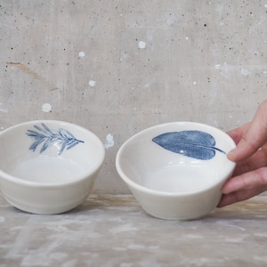 Puede incluir: Dos cuencos de cerámica blanca con diseños de hojas azules. El cuenco de la izquierda tiene una rama de hojas, mientras que el cuenco de la derecha tiene una sola hoja grande.