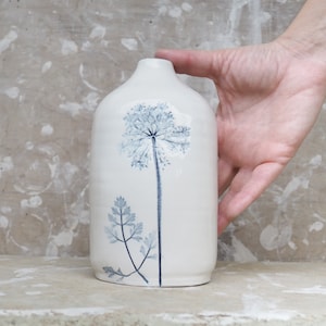 Op de afbeelding: Witte keramische vaas met een blauw bloemenontwerp. De vaas heeft een hoge, delicate bloem met een tros kleine bloesems aan de bovenkant en een kleinere, bladerige stengel aan de onderkant.