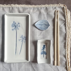 Puede incluir: Un juego de cuatro platos de cerámica blanca con diseños florales azules. Los platos incluyen una fuente rectangular, un plato rectangular pequeño, un plato con forma de hoja y un par de palillos.