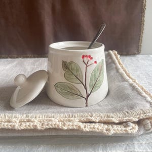 Puede incluir: Un azucarero de cerámica blanca con tapa y una cucharita. El cuenco está decorado con un diseño floral verde y rojo. La tapa está a un lado. El cuenco está sobre un salvamanteles blanco y beige de ganchillo.