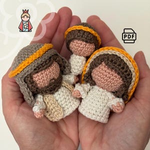 May include: Three crocheted figures of religious figures are held in open hands. The figures have brown hair, tan skin, and are wearing white and tan clothing. The figures are crocheted with brown, tan, and yellow yarn. The figures are small and appear to be handmade.