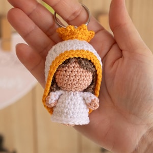 Puede incluir: Un llavero de ganchillo con una pequeña muñeca con un vestido blanco, una corona amarilla y cabello castaño. La muñeca lleva un vestido blanco con ribete amarillo y una corona amarilla.