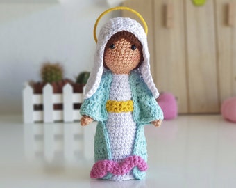 May include: A crocheted doll of the Virgin Mary, wearing a blue and white dress with a yellow sash, and a white head covering. She has a halo above her head and brown hair.