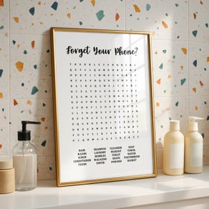 A gold-framed print with the title "Forget Your Phone?" and a word search puzzle with bathroom-related words, standing on a white counter against a colorful tiled wall.