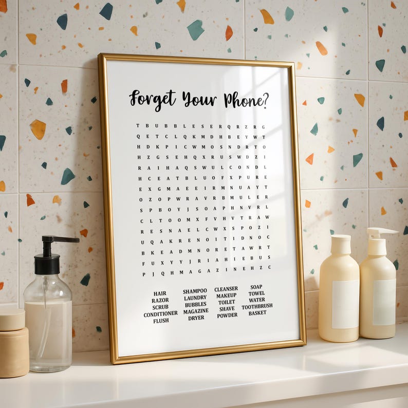 A gold-framed print with the title "Forget Your Phone?" and a word search puzzle with bathroom-related words, standing on a white counter against a colorful tiled wall.