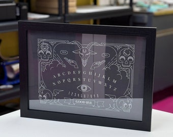 May include: A black framed spirit board print featuring white line art of a goat head, skulls, hands, an eye, and the alphabet with numbers 1-9, 0, and the text YES, NO, and GOOD BYE. The design includes celestial symbols and decorative clouds.