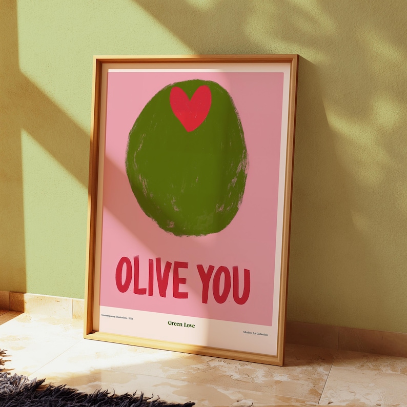 May include: A framed art print featuring a green olive with a red heart on a pink background. The words "OLIVE YOU" are in red below the olive. The print is in a wooden frame and leans against a green wall.
