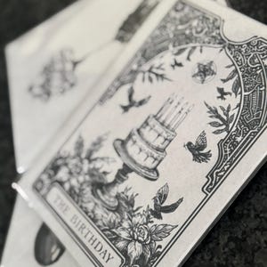 Puede incluir: Una pila de tarjetas de felicitaci&oacute;n con una ilustraci&oacute;n en blanco y negro. La tarjeta presenta una tarta de cumplea&ntilde;os de varios niveles con velas encendidas, p&aacute;jaros y elementos florales. El texto "THE BIRTHAY" est&aacute; en la parte inferior de la tarjeta.