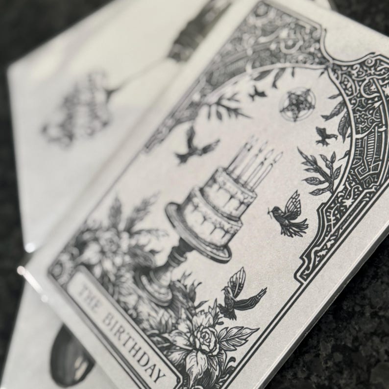 Puede incluir: Una pila de tarjetas de felicitaci&oacute;n con una ilustraci&oacute;n en blanco y negro. La tarjeta presenta una tarta de cumplea&ntilde;os de varios niveles con velas encendidas, p&aacute;jaros y elementos florales. El texto "THE BIRTHAY" est&aacute; en la parte inferior de la tarjeta.