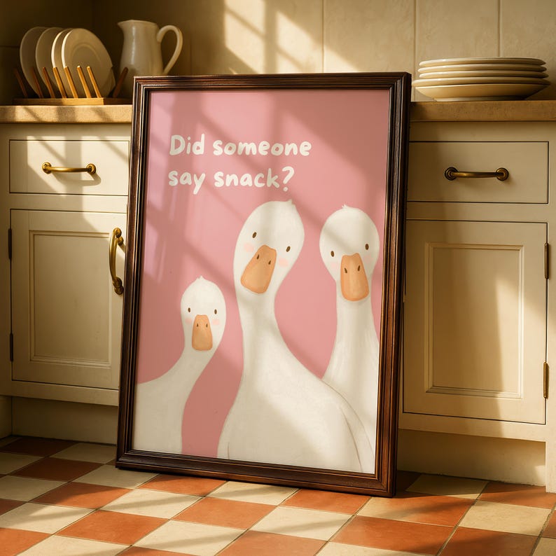 A framed print with a dark wood border features three white geese on a pink background, with the text "Did someone say snack?" in white, leaning against kitchen cabinets.
