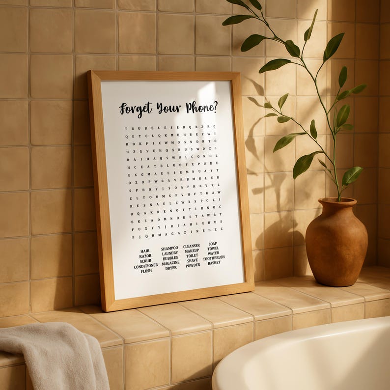 A light wood-framed print with "Forget Your Phone?" and a word search puzzle, leaning against a tiled wall on the edge of a bathtub, with a potted plant nearby.
