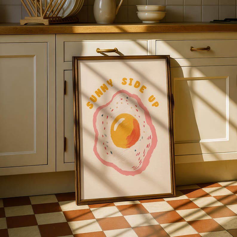A framed print of a sunny-side-up egg with a pink and yellow border and the words "SUNNY SIDE UP" in yellow letters, leaning against light green kitchen cabinets on a checkered floor.