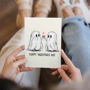 Puede incluir: Tarjeta de San Valentín con una ilustración en blanco y negro de dos fantasmas sosteniendo un corazón rojo. El texto "HAPPY VALENTINES DAY" está impreso debajo de la imagen de los fantasmas.