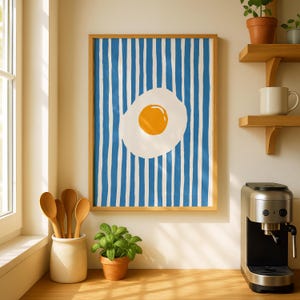 May include: A framed wall art print featuring a centered sunny-side up egg with a bright orange yolk and white egg white, set against a background of vertical blue and white stripes. The frame is a light-toned wood, displaying a clean aesthetic.