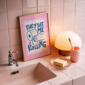 A framed print with a pink border, a light pink background, and the words "THEY SEE ME ROLLING" in blue, with a hand holding a toilet paper roll, sits on a pink tiled bathroom counter next to a sink, soap dish, toothbrush, and lamp.