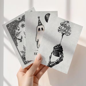 Puede incluir: Tres tarjetas de felicitaci&oacute;n en escala de grises se sostienen en una mano. Una tarjeta presenta un dise&ntilde;o floral y de aves detallado. Otra muestra un fantasma con un gorro de fiesta y un globo. La tercera tarjeta tiene una magdalena sostenida por una mano de esqueleto con el texto "Happy Birthday".