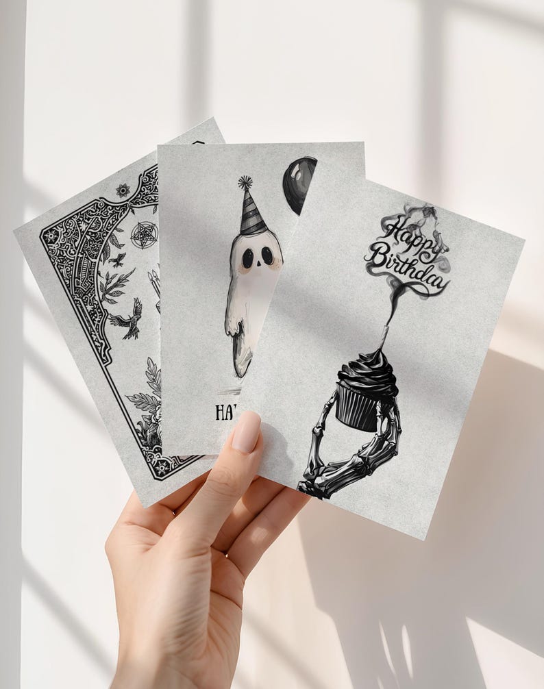 Puede incluir: Tres tarjetas de felicitaci&oacute;n en escala de grises se sostienen en una mano. Una tarjeta presenta un dise&ntilde;o floral y de aves detallado. Otra muestra un fantasma con un gorro de fiesta y un globo. La tercera tarjeta tiene una magdalena sostenida por una mano de esqueleto con el texto "Happy Birthday".