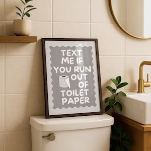 May include: A framed rectangular wall art piece with a gray background and a scalloped white border. The text reads: TEXT ME IF YOU RUN OUT OF TOILET PAPER, accompanied by a simple line drawing of a roll of toilet paper in the center of the print.