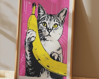 May include: A framed wall art print featuring a black and white tabby cat with yellow eyes holding a large yellow banana against a vibrant pink background. The graphic illustration has a distressed, textured appearance within a light wood frame.