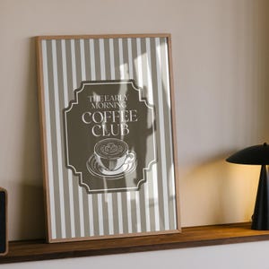 May include: Framed print featuring a coffee-themed design. The artwork has a brown and white striped background with a central label that reads "THE EARLY MORNING COFFEE CLUB." The label includes a coffee cup illustration. The frame is made of wood.