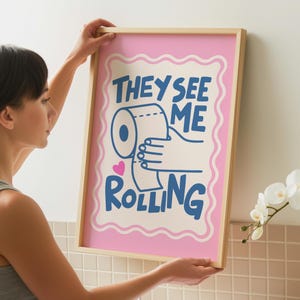 A person holds up a framed print with a pink border, a light pink background, and the words "THEY SEE ME ROLLING" in blue, with a hand holding a toilet paper roll.