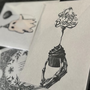 Puede incluir: Tarjetas de felicitaci&oacute;n con ilustraciones &uacute;nicas. Una tarjeta presenta una magdalena sostenida por una mano esquel&eacute;tica, con las palabras "Happy Birthday" arriba. Otra tarjeta muestra un fantasma con un gorro de fiesta y un globo. Las tarjetas son en blanco y negro.