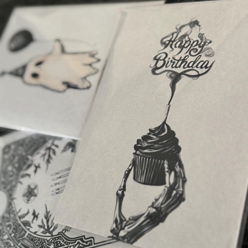 Puede incluir: Tarjetas de felicitaci&oacute;n con ilustraciones &uacute;nicas. Una tarjeta presenta una magdalena sostenida por una mano esquel&eacute;tica, con las palabras "Happy Birthday" arriba. Otra tarjeta muestra un fantasma con un gorro de fiesta y un globo. Las tarjetas son en blanco y negro.