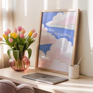 May include: A wooden framed art print with the text "Busy Dilly Dallying" features a blue sky and pink and white clouds. A vase of pink and yellow tulips sits next to a laptop and a speckled ceramic pen holder.