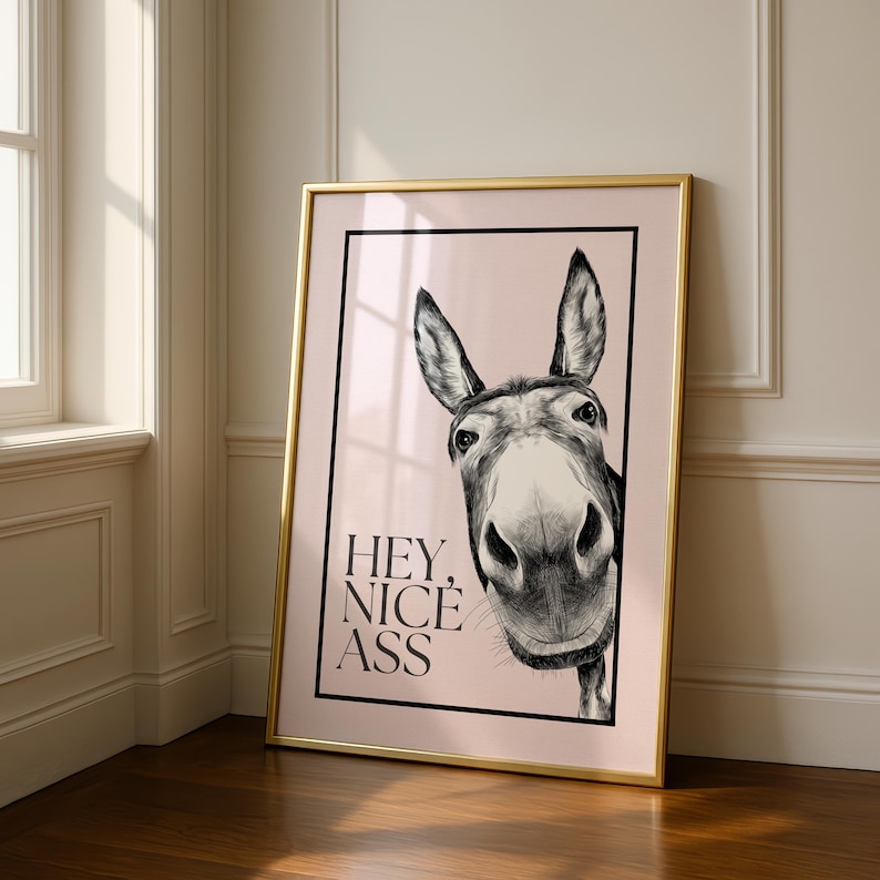 May include: Framed art print featuring a detailed black and white illustration of a donkey's face. The donkey is centered, with the text "HEY, NICE ASS" below. The print is set against a light pink background, framed in gold.