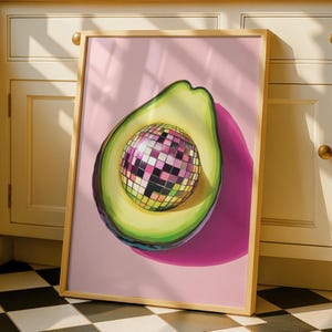 May include: A framed art print featuring a whimsical avocado design. The avocado is cut open, revealing a disco ball center. The background is a soft pink, and the frame is a light wood color. The artwork is displayed on a black and white checkered floor.