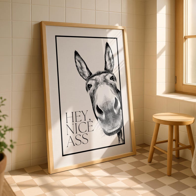 May include: Framed black and white donkey art print. The donkey's face is centered, with the text "HEY, NICE ASS" below. The print is in a light wood frame and leans against a tiled wall.
