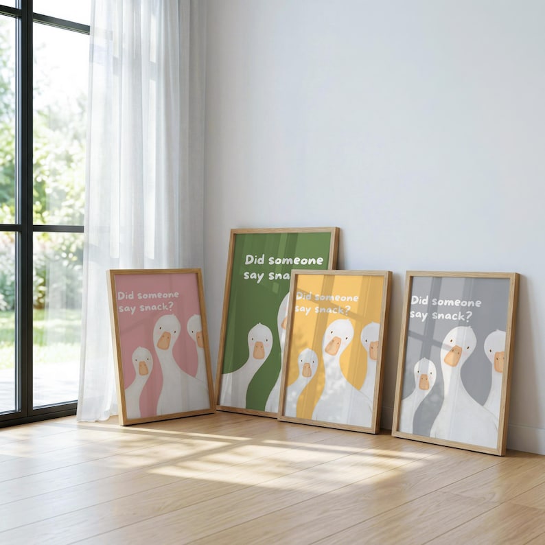 Four framed prints lean against a white wall, each featuring three white geese and the text "Did someone say snack?" on backgrounds of pink, green, yellow, and gray.