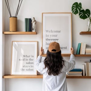 May include: Two framed wall art prints with text. One print reads Welcome to your therapy appointment and the other reads What is said in the salon stays in the salon. The prints are displayed on wooden shelves alongside books and a small statue.