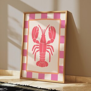 A framed print of a pink lobster on a cream background with a pink and white checkered border, leaning against a light-colored wall on a wooden floor.