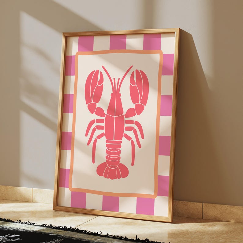 A framed print of a pink lobster on a cream background with a pink and white checkered border, leaning against a light-colored wall on a wooden floor.