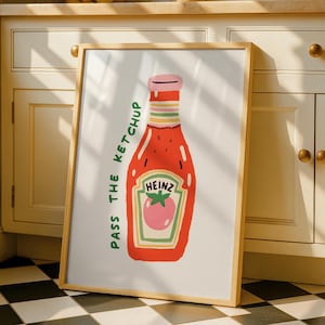 Op de afbeelding: Een ingelijste print met een gestileerde illustratie van een ketchupfles. De fles is rood met een roze dop en een label met "HEINZ" en een tomaat. De tekst "PASS THE KETCHUP" staat verticaal.