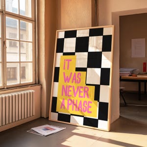 Può includere: Una stampa incorniciata con uno sfondo a scacchi bianco e nero. Il testo "IT WAS NEVER A PHASE" è scritto in lettere rosa e gialle su uno sfondo giallo.