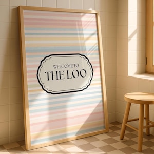 Puede incluir: Un póster enmarcado con un marco de madera clara. El póster presenta rayas horizontales en colores pastel y el texto "WELCOME TO THE LOO" en una fuente negra decorativa dentro de una forma blanca y negra. El póster está apoyado contra una pared.