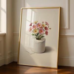 Puede incluir: Impresión artística enmarcada que presenta margaritas rosas y amarillas en un jarrón de rollo de papel higiénico blanco. La obra de arte tiene un marco dorado y está sobre un fondo neutro.