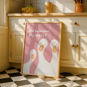 A framed print with a light wood border features three white geese on a pink background, with the text "Did someone say snack?" in white, leaning against kitchen cabinets.
