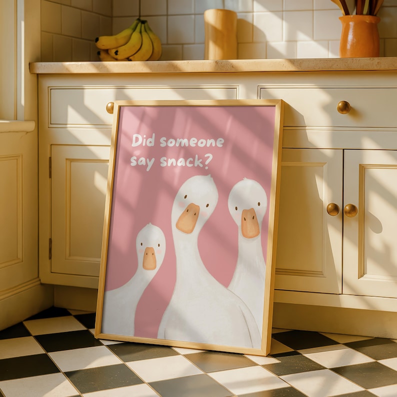A framed print with a light wood border features three white geese on a pink background, with the text "Did someone say snack?" in white, leaning against kitchen cabinets.