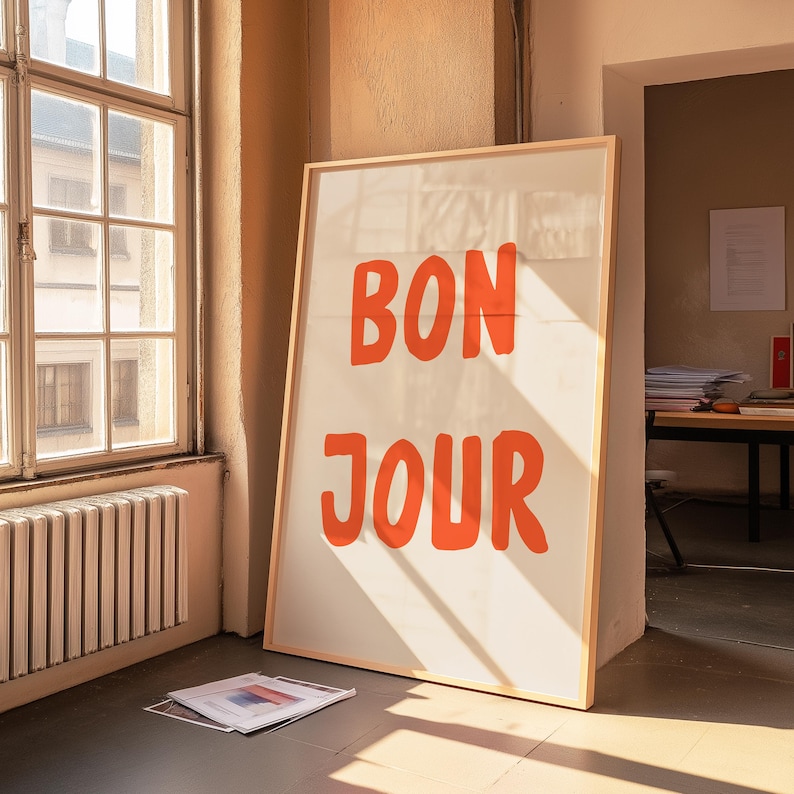 May include: A large framed print with the words "BON JOUR" in bold orange letters. The print is leaning against a wall in a room with a window and sunlight streaming in. A few papers are on the floor.