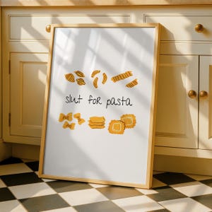 May include: A framed white print with a wooden frame. The print features various pasta shapes in yellow and orange, with the phrase "slut for pasta" written in black script. The background is a kitchen setting.