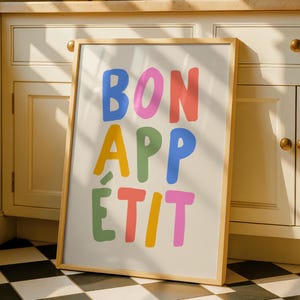 May include: A framed art print with the words "BON APPÉTIT" in colourful, bold letters. The letters are in blue, pink, red, yellow, green, and orange. The print has a light wood frame and is leaning against a white cabinet.
