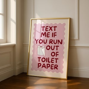 May include: A framed wall art print featuring a light pink scalloped rectangle background with dark red text that reads "TEXT ME IF YOU RUN OUT OF TOILET PAPER" and a simple line drawing of a white toilet paper roll centered in the middle.