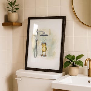 May include: A framed art print featuring a whimsical illustration of a frog taking a shower. The frog, with a shower cap, stands under a silver shower head. The artwork is set against a soft, watercolor-style background, framed in a dark brown frame.