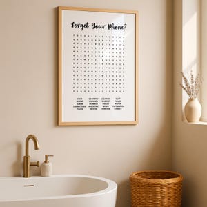 A light wood-framed print with "Forget Your Phone?" and a word search puzzle, resting on a bathroom counter next to a white sink and a gold faucet.