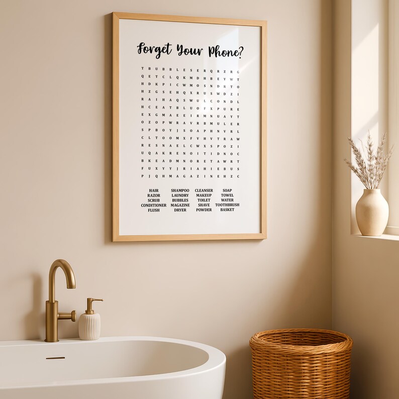 A light wood-framed print with "Forget Your Phone?" and a word search puzzle, resting on a bathroom counter next to a white sink and a gold faucet.
