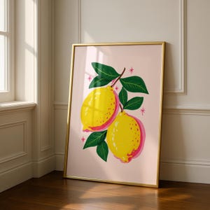 A gold framed print of two yellow and pink lemons on a branch with green leaves, on a light pink background with small pink starbursts, leaning against a white wall and wood floor.
